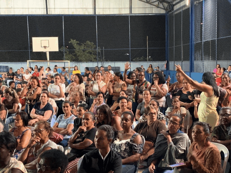 Atingidas de Barra Longa estão em reunião num ginásio. Algumas estão com as mãos levantadas, enquanto uma delas diz algo.