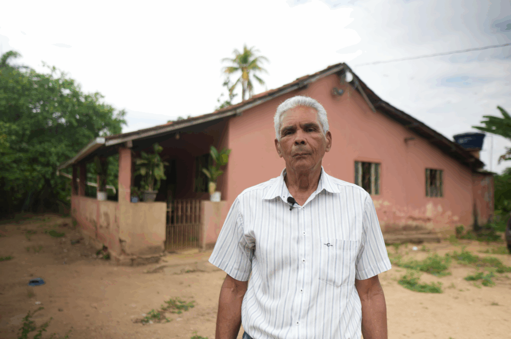 Geraldo Magela é um senhor grisalho, que olha para a câmera em frente a uma casa de cor salmão. Entre ele e a casa há um pátio descampado. O céu está nublado e acima da casa pode-se ver um palmeira bem alta.