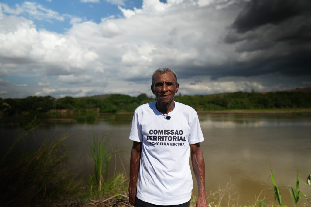 Seu Itamar é um homem negro, idoso, vestindo uma camiseta com os dizeres "Comissão Territorial Cachoeira Escura". Ele está de frente para a câmera, com o Rio Doce às suas costas.