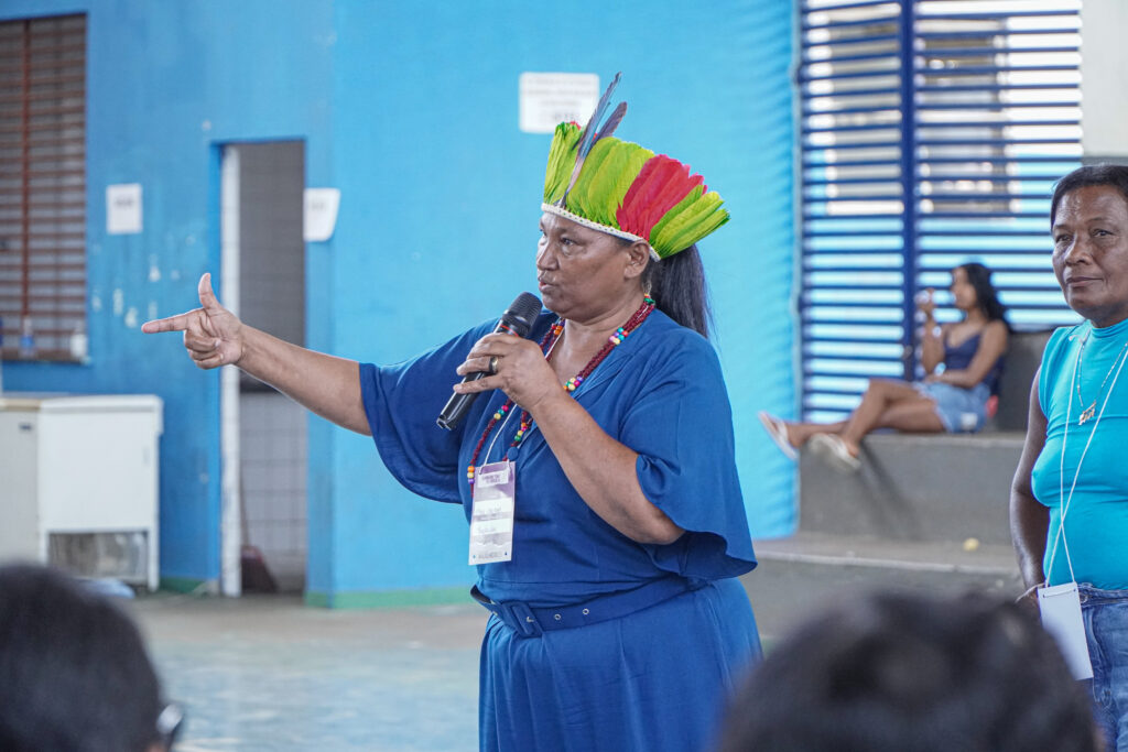 Maru Orutum Puri, a Maria José, é uma liderança indígena. Na foto, ela aponta para o lado esquerdo da tela. Ela usa um vestido azul com cinto da mesma cor, e adereços indígenas ao redor do pescoço. Na cabeça, usa um cocar de penas verdes e vermelhas, com penas azuis no centro. Ela usa um crachá, e segura um microfone. Atrás dela, um espaço com paredes e grades azuis.