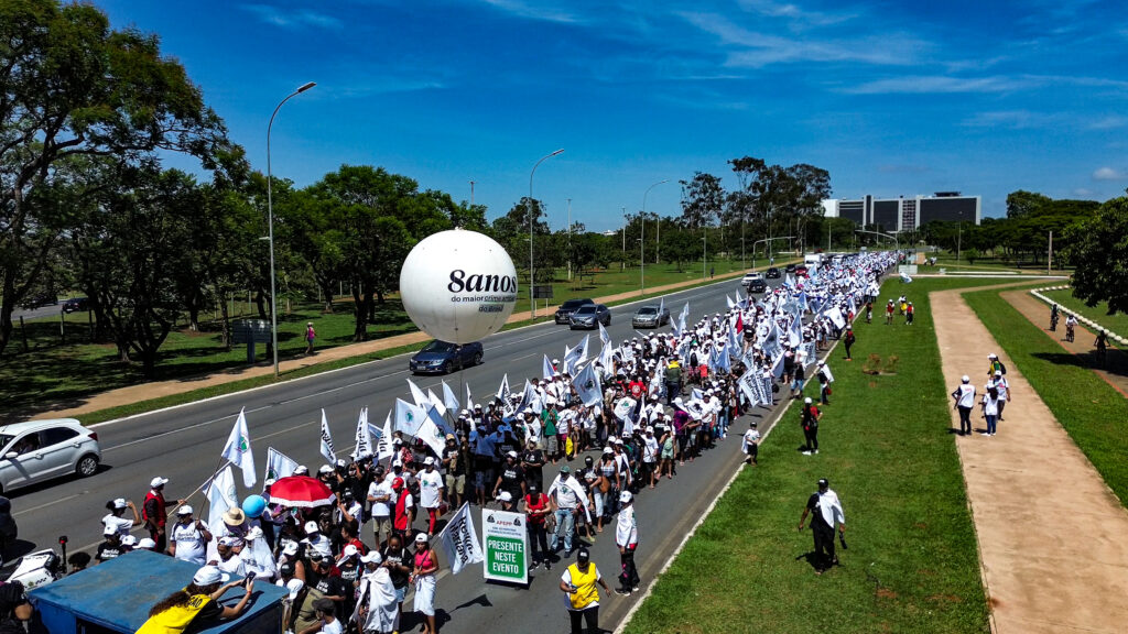 Atingidos fazem passeata em Brasília, ocupando uma faixa de uma extensa avenida. Eles seguram bandeiras e há um balão escrito "8 anos", o que situa a passeata no final de 2023. Eles estão cercados por áreas verdes e o céu é bem azulado.