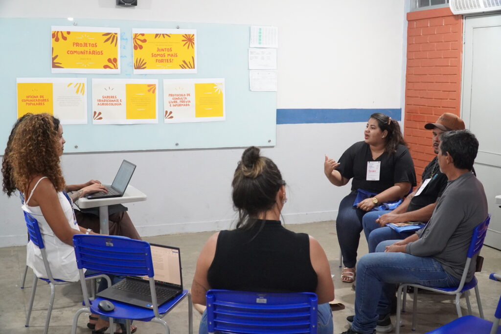 Atingidos e atingidas participam de formação sobre Projetos Comunitários. Estão em uma sala de escola municipal, com cadeiras azuis. No quadro da sala, cartazes com os escritos: "projetos comunitários", "juntos somos mais fortes", "oficina de comunicadores populares", "Saberes tradicionais e agroecologia" e "protocolo de consulte livre, prévia e informada". Esses cartazes, de cor amarela e fonte avermelhada, possuem mais texto mas são ilegíveis pela distância. À esquerda da foto, duas técnicas da Aedas escutam a fala da atingida que está à direita, juntamente com dois outros atingidos. Ao centro, uma sexta pessoa, provavelmente da Assessoria, escuta o relato da atingida.