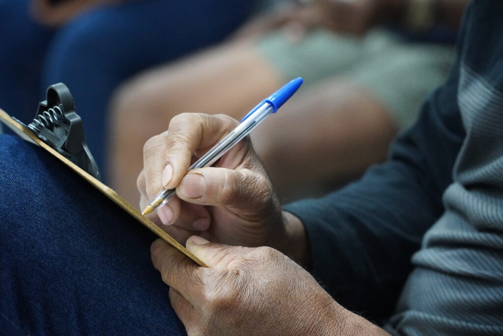 Foto das mãos de um atingido em close-up preenchendo algo em uma prancheta. Não conseguimos ver o conteúdo da prancheta. A caneta é uma esferográfica de tampa azul. O atingido sustenta a prancheta em sua perna.